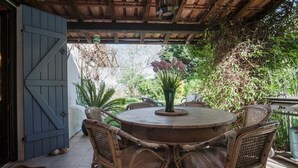 A covered patio with a round table and wicker chairs, surrounded by greenery and a blue door.