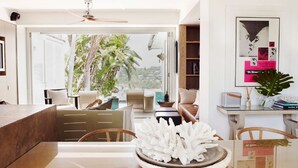 A modern living room with a large window, a dining area with a white centerpiece, and a bookshelf with books and decorative items. - The Elysium by Contemporary Hotels (Palm Beach)