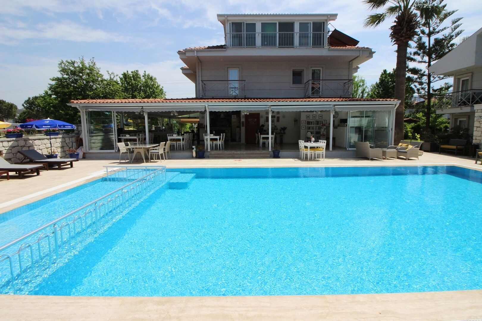 A swimming pool with a covered patio area, lounge chairs, and a two-story building in the background.