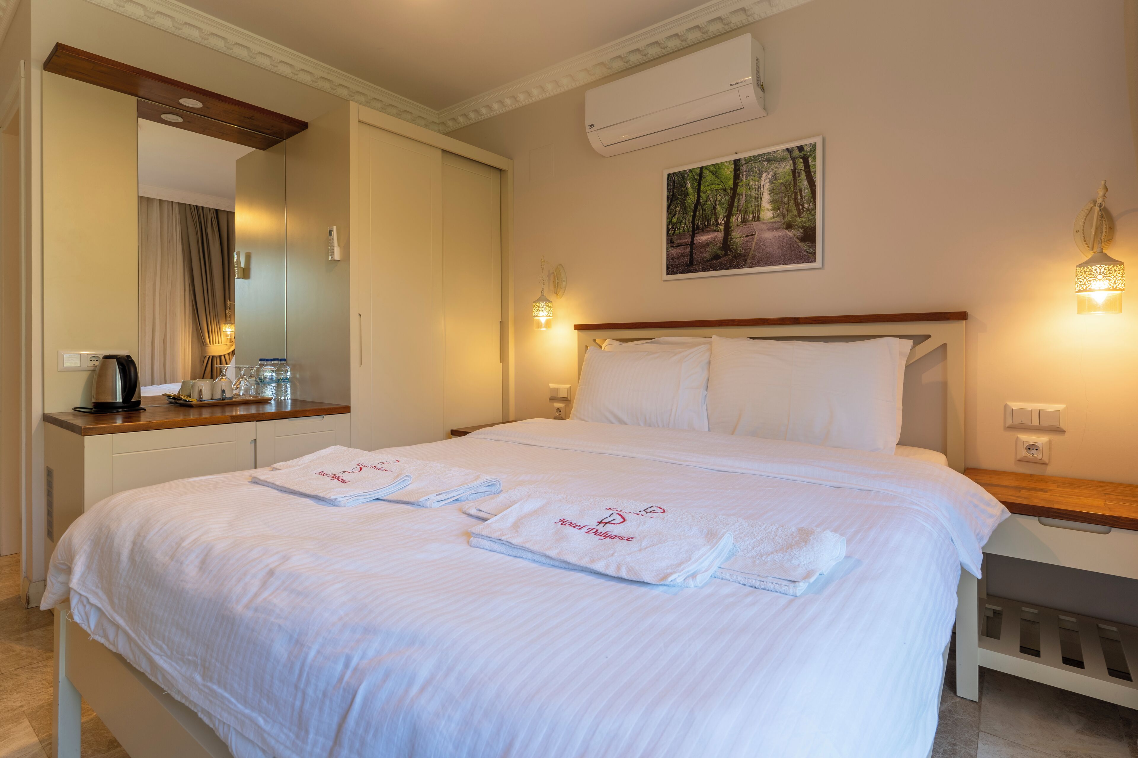 A neatly made bed with white linens and towels, a bedside table with a lamp, and a wall-mounted air conditioner.