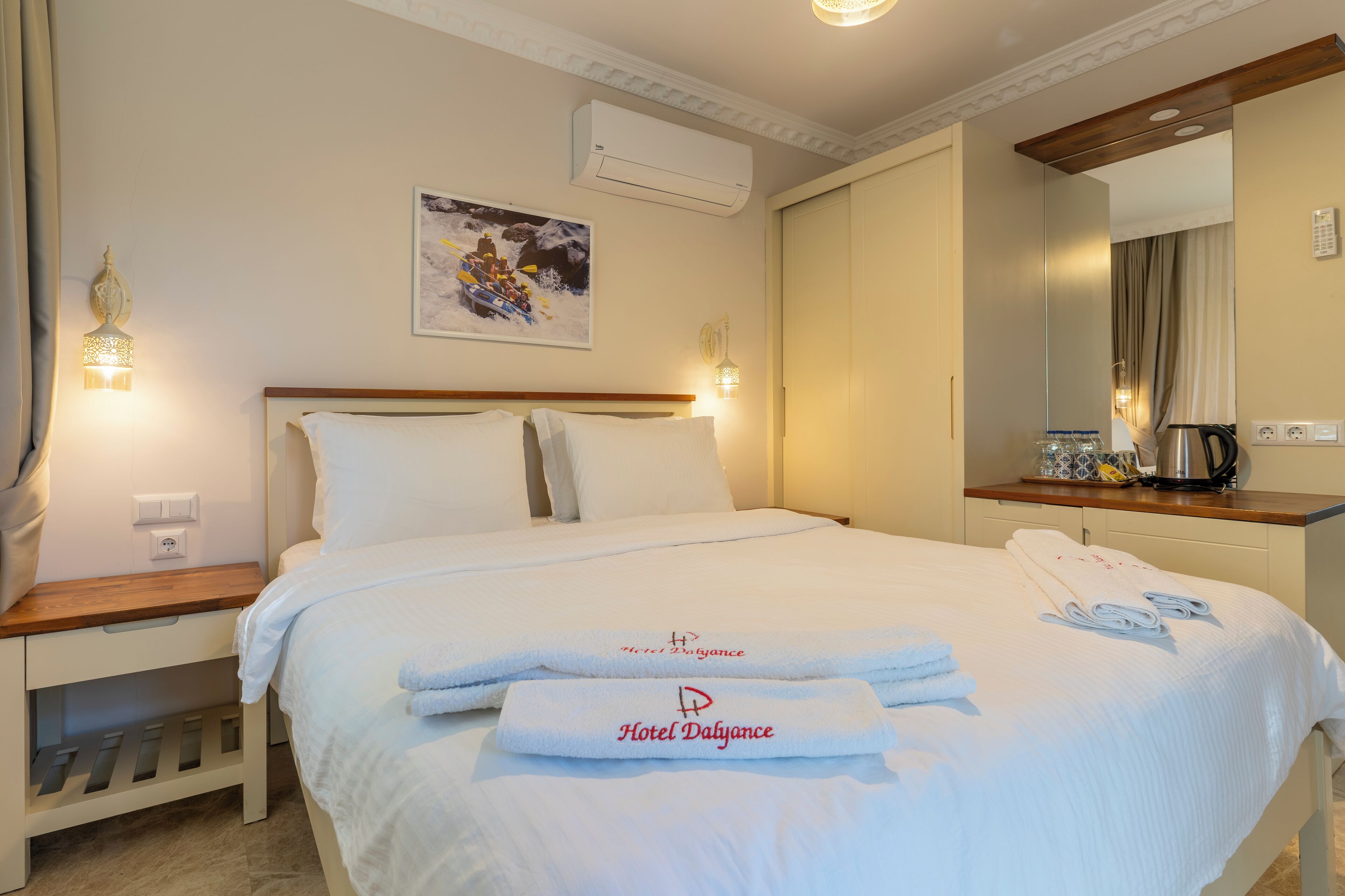 A hotel room with a neatly made bed, a bedside table with a lamp, a wall-mounted air conditioner, and a painting above the bed.