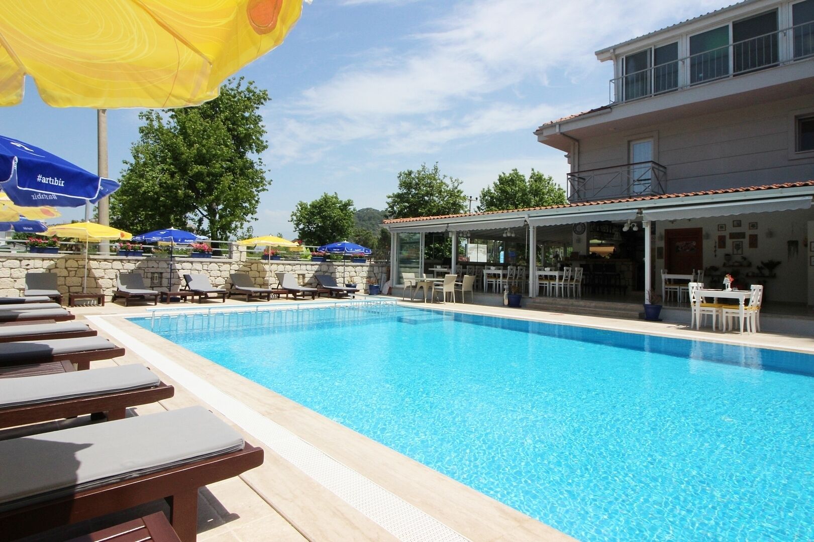A swimming pool with lounge chairs and umbrellas, a covered outdoor seating area, and a building with a tiled roof.