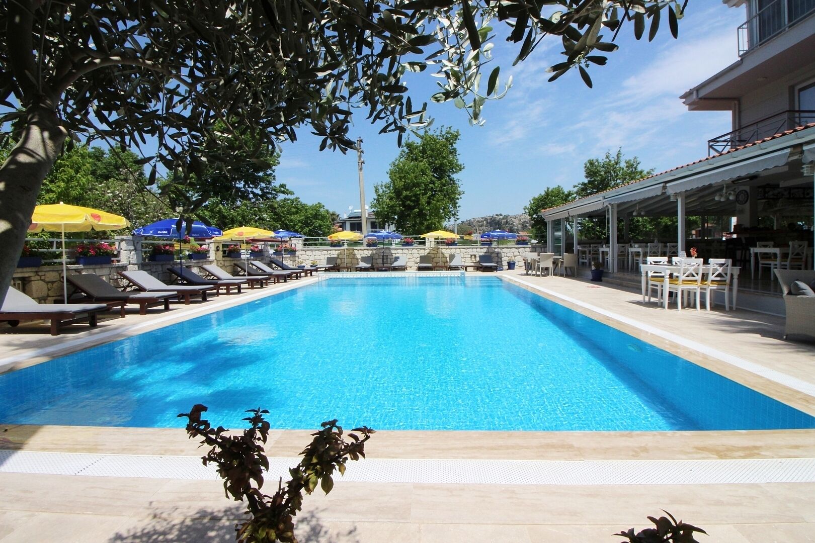 A rectangular swimming pool with clear blue water, surrounded by lounge chairs and umbrellas, and a covered seating area with tables and chairs.