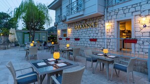 Un espace extérieur aménagé avec des meubles et des tables en osier, un bâtiment en pierre dont la façade est illuminée et sur laquelle on peut lire le nom « BALYANCE », et un arbre qui offre de l’ombre.