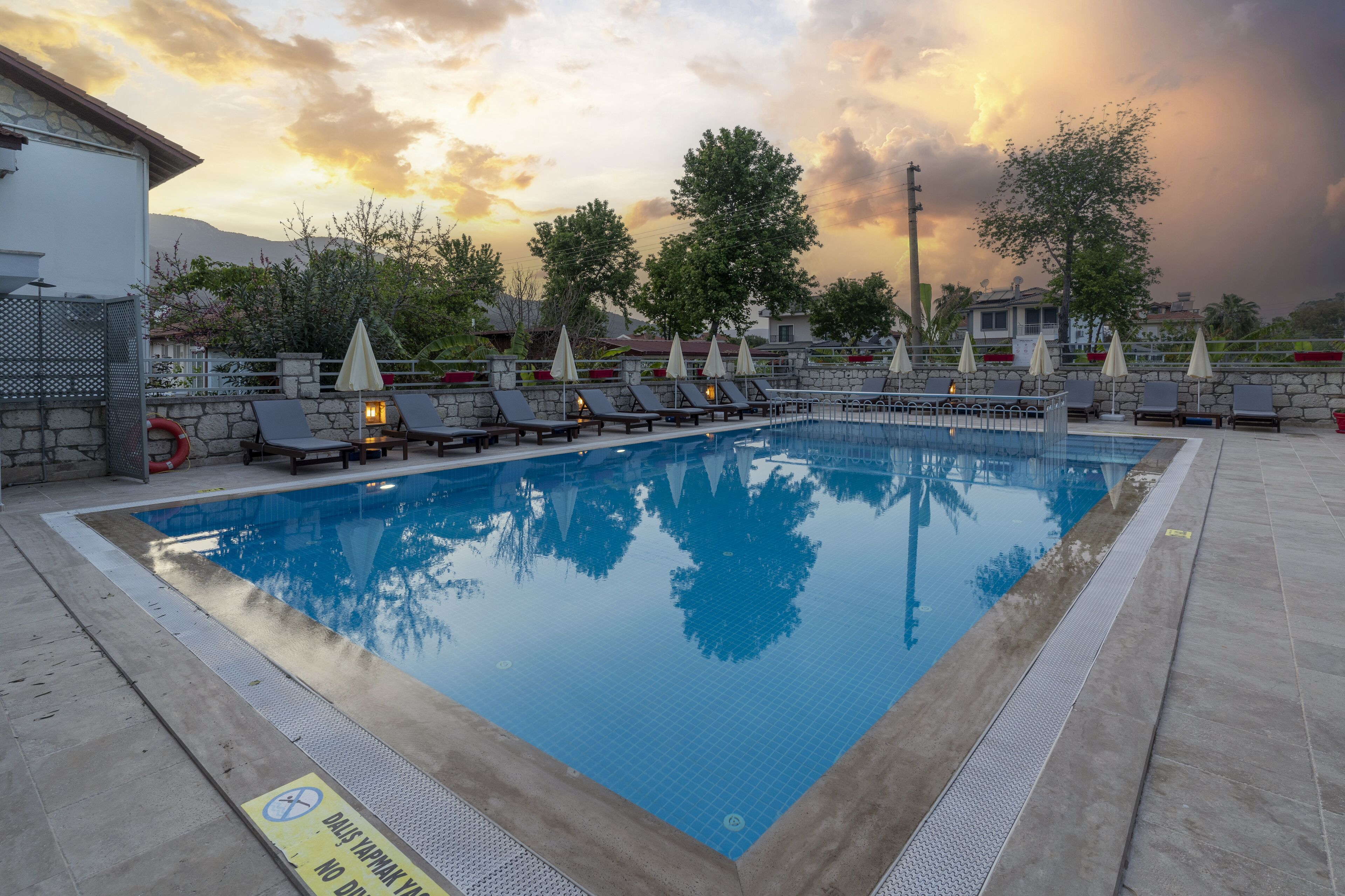 Une piscine entourée de chaises longues et de parasols, bordée de murs de pierre et d’arbres, avec un coucher de soleil en arrière-plan.