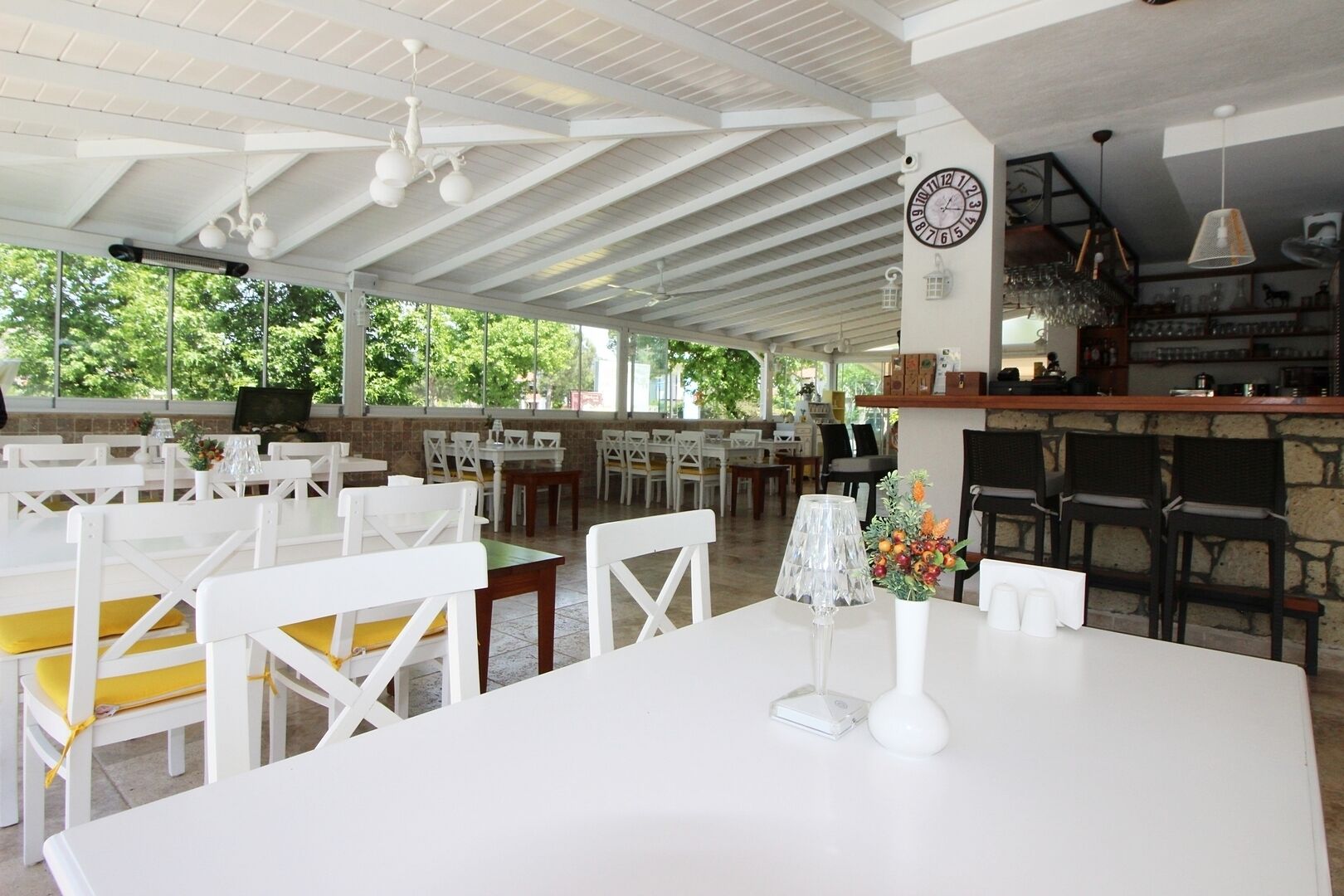 An outdoor dining area with white tables and chairs, a bar area with a clock, and a view of greenery outside.