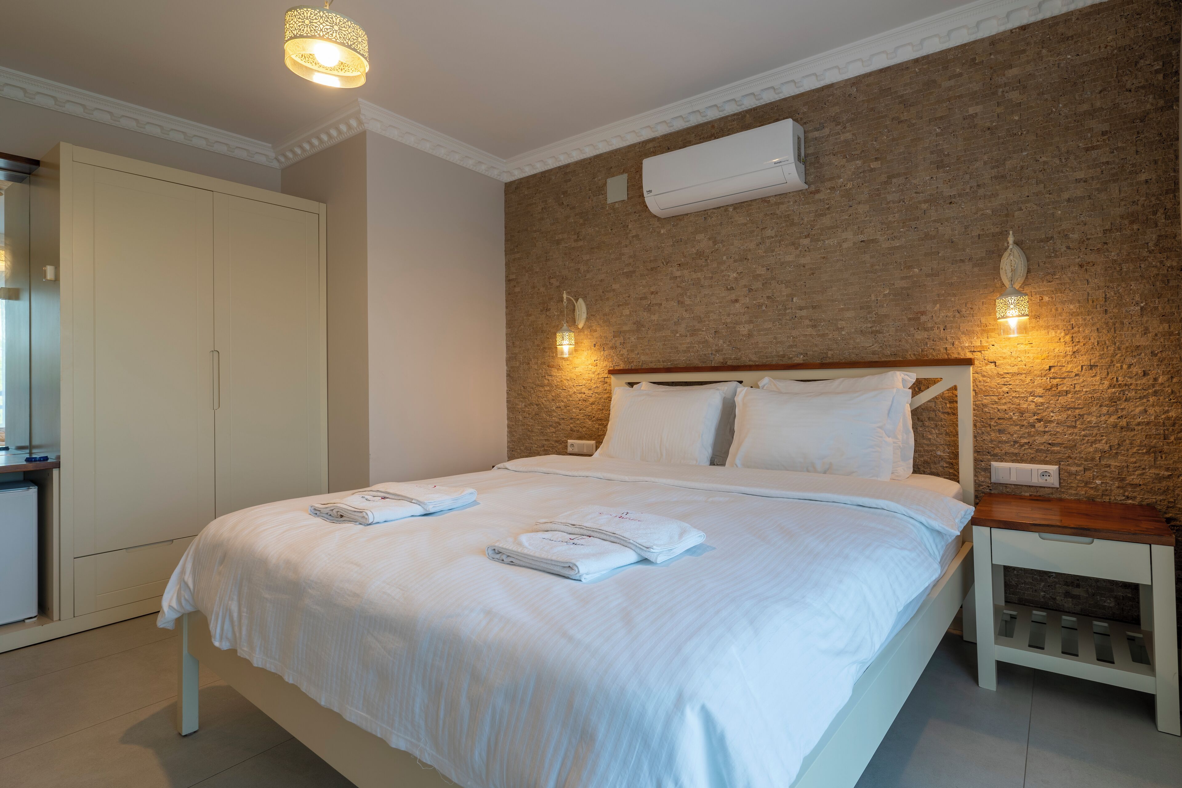Ein ordentlich bezogenes Bett mit weißen Leinen und Handtüchern, ein hölzernes Kopfende und eine Wand mit Ziegeloptik mit zwei Wandleuchten.