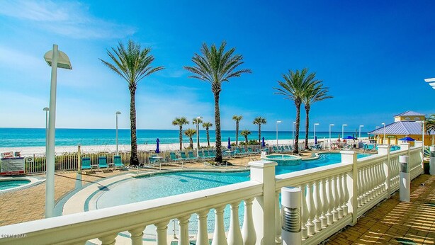 Zona de piscina en un resort, con palmeras, una piscina y una playa al fondo.