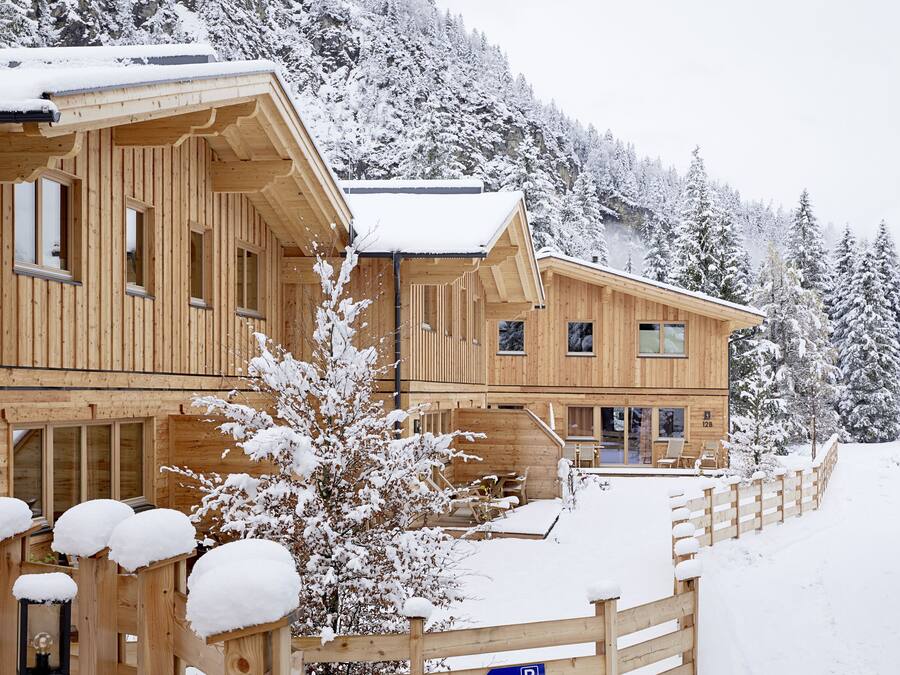 Una cabaña de madera con múltiples ventanas y un balcón, rodeada de árboles cubiertos de nieve y un paisaje montañoso.