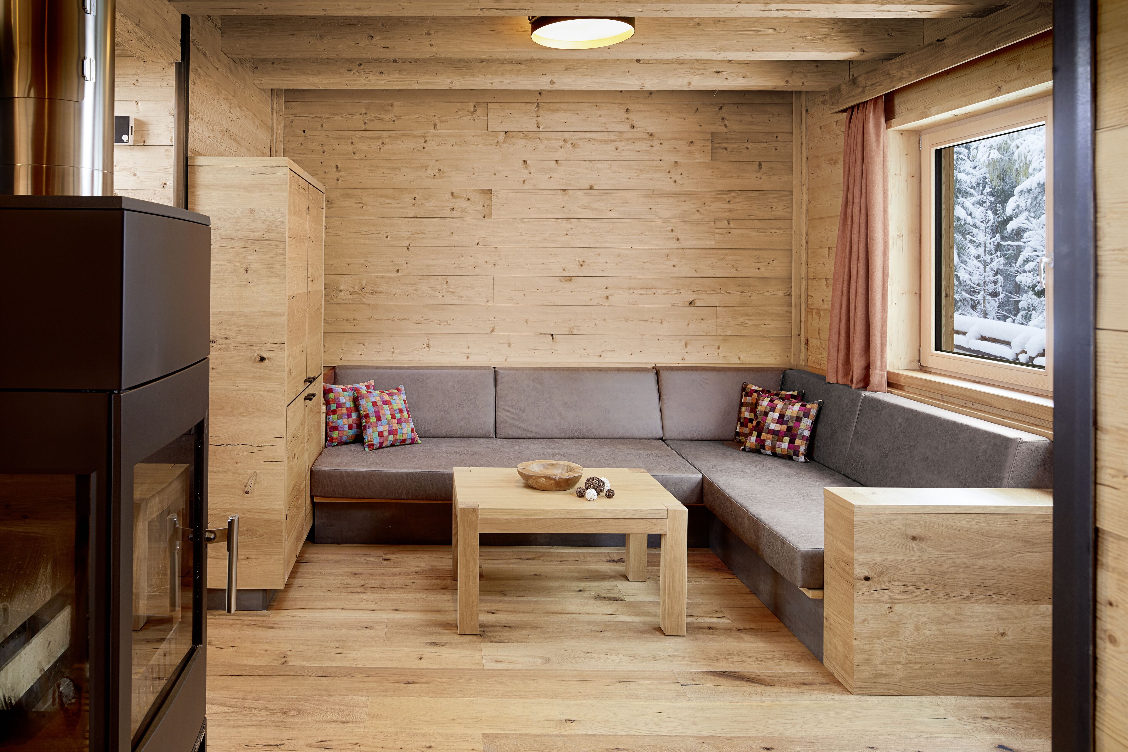 A cozy cabin interior with a wooden floor, a sofa with cushions, a small table, and a window with a view of snow-covered trees.