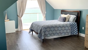 A bedroom with a bed, a nightstand, a window with curtains, and a view of the sea.