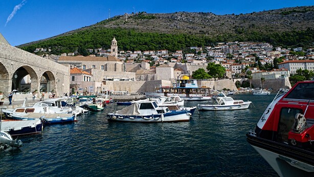 A harbor with boats docked, a historic stone bridge, and a town with buildings and a hill in the background. - Old Town Center Studio Apartments (Dubrovnik)