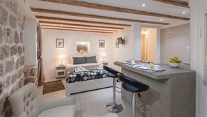 A modern bedroom with a stone wall, a bed with a patterned bedspread, a nightstand with a lamp, and a breakfast bar with fruit. - Old Town Center Studio Apartments (Dubrovnik)