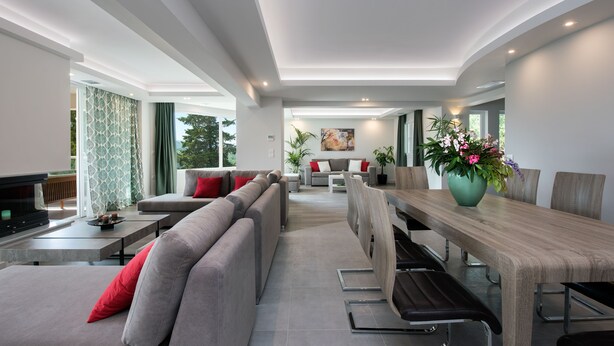 A modern living room with a sofa, dining table, and chairs. There is a fireplace, a large window, and a painting on the wall. - Gem Villa in Viros Corfu  (Corfu)