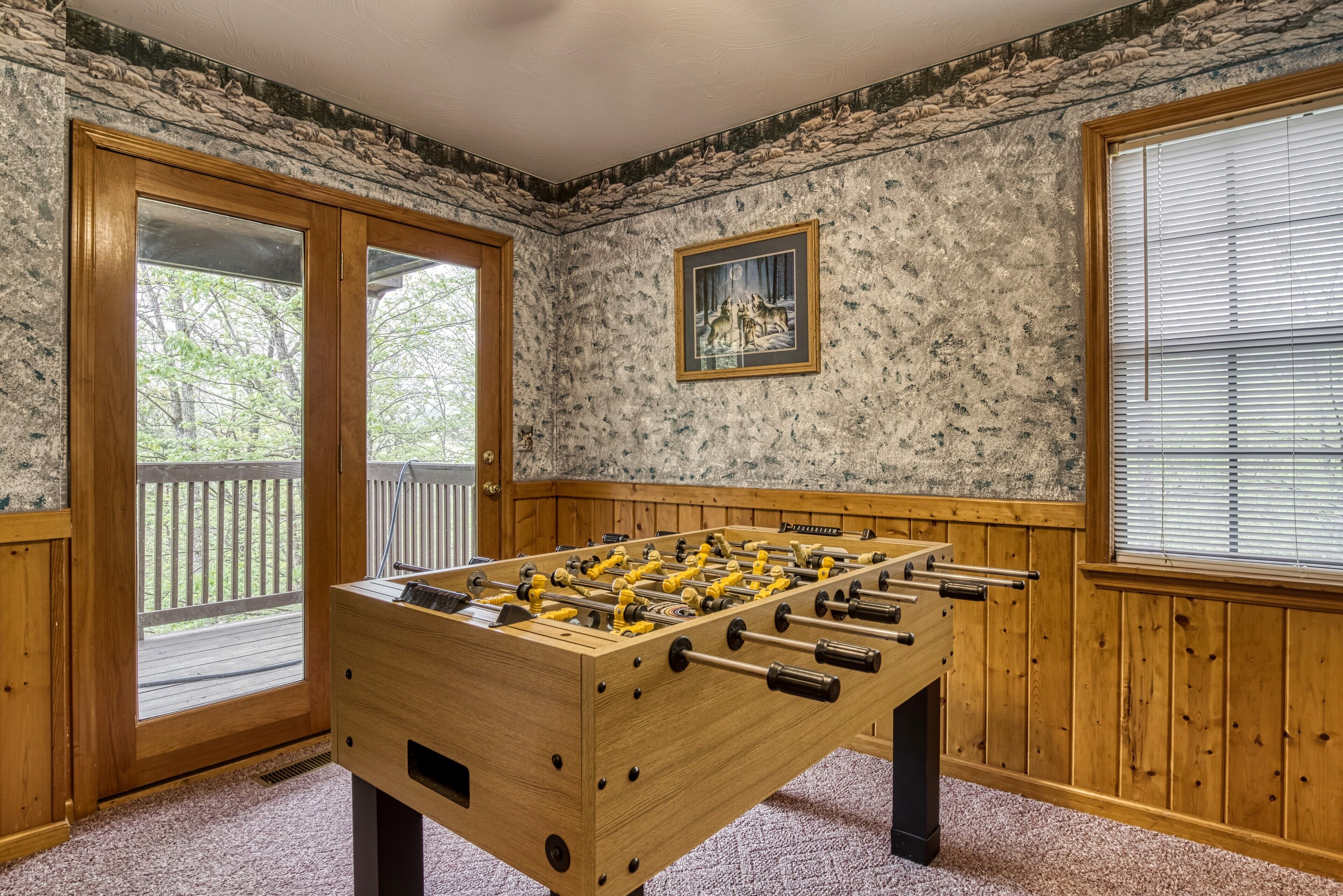 A wooden foosball table in a room with stone-patterned walls, a framed picture, and a wooden door leading to an outdoor deck.