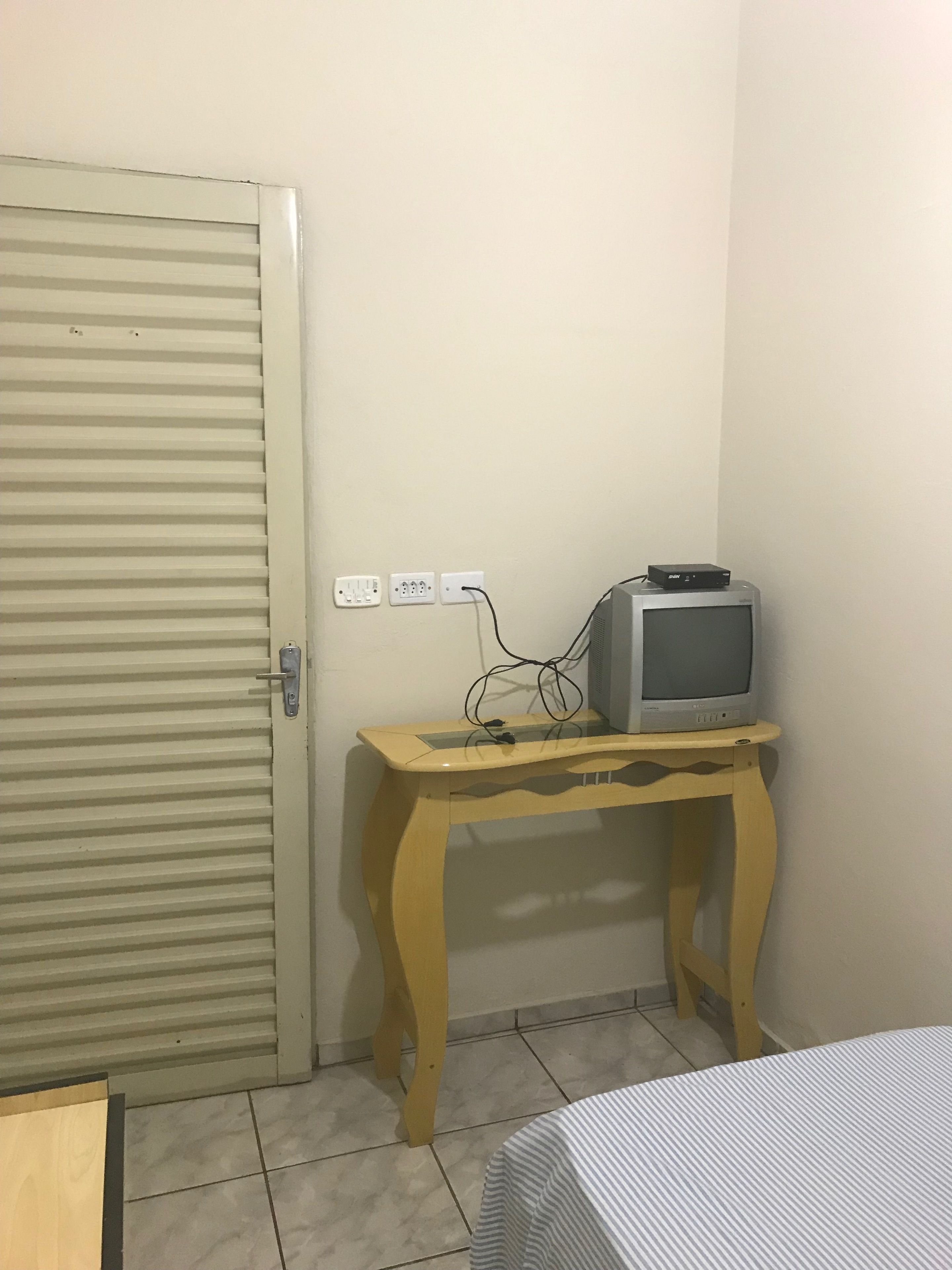 Um quarto com porta fechada, uma mesa de madeira com uma televisão em cima e uma cama com colcha listrada.