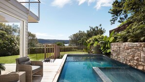 Une piscine entourée d’une terrasse en bois, de meubles en osier et d’un mur en pierre.
