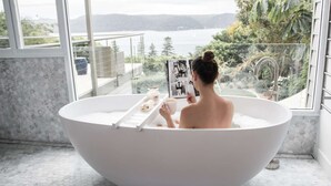 Une personne est assise dans une grande baignoire blanche, en train de lire un magazine, et on aperçoit, à travers de grandes fenêtres, un lac et des montagnes.