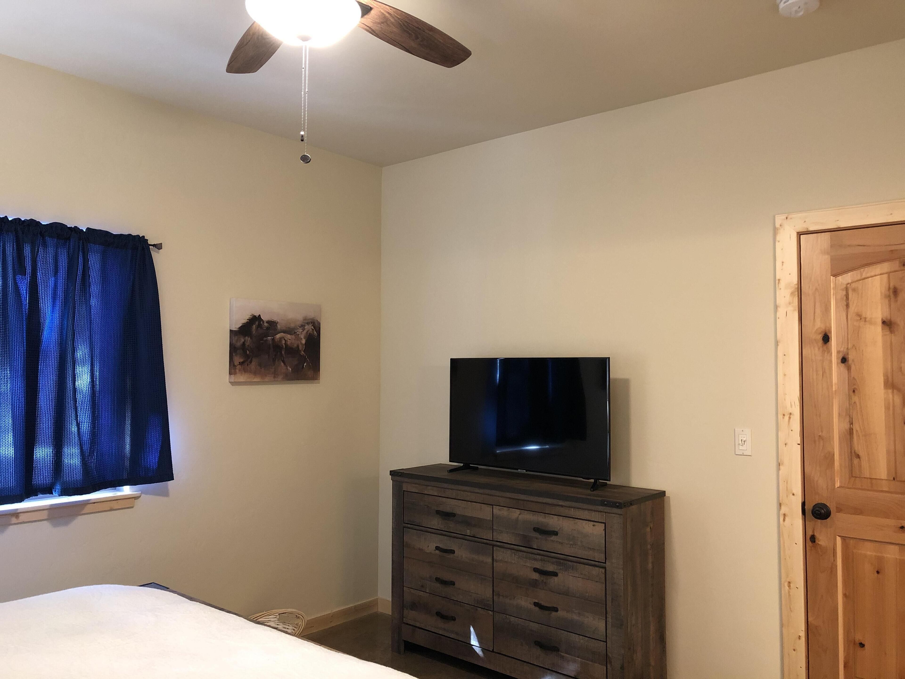 Une chambre à coucher comprenant un lit, une commode, un téléviseur, un ventilateur de plafond, une fenêtre avec des rideaux et une porte.