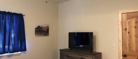 Une chambre à coucher comprenant un lit, une commode, un téléviseur, un ventilateur de plafond, une fenêtre avec des rideaux et une porte.