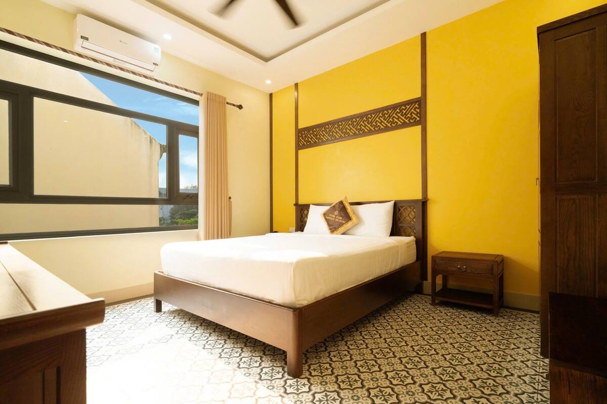 Quarto de hotel com uma cama, uma pequena mesa de cabeceira, uma janela com cortinas e um piso estampado.