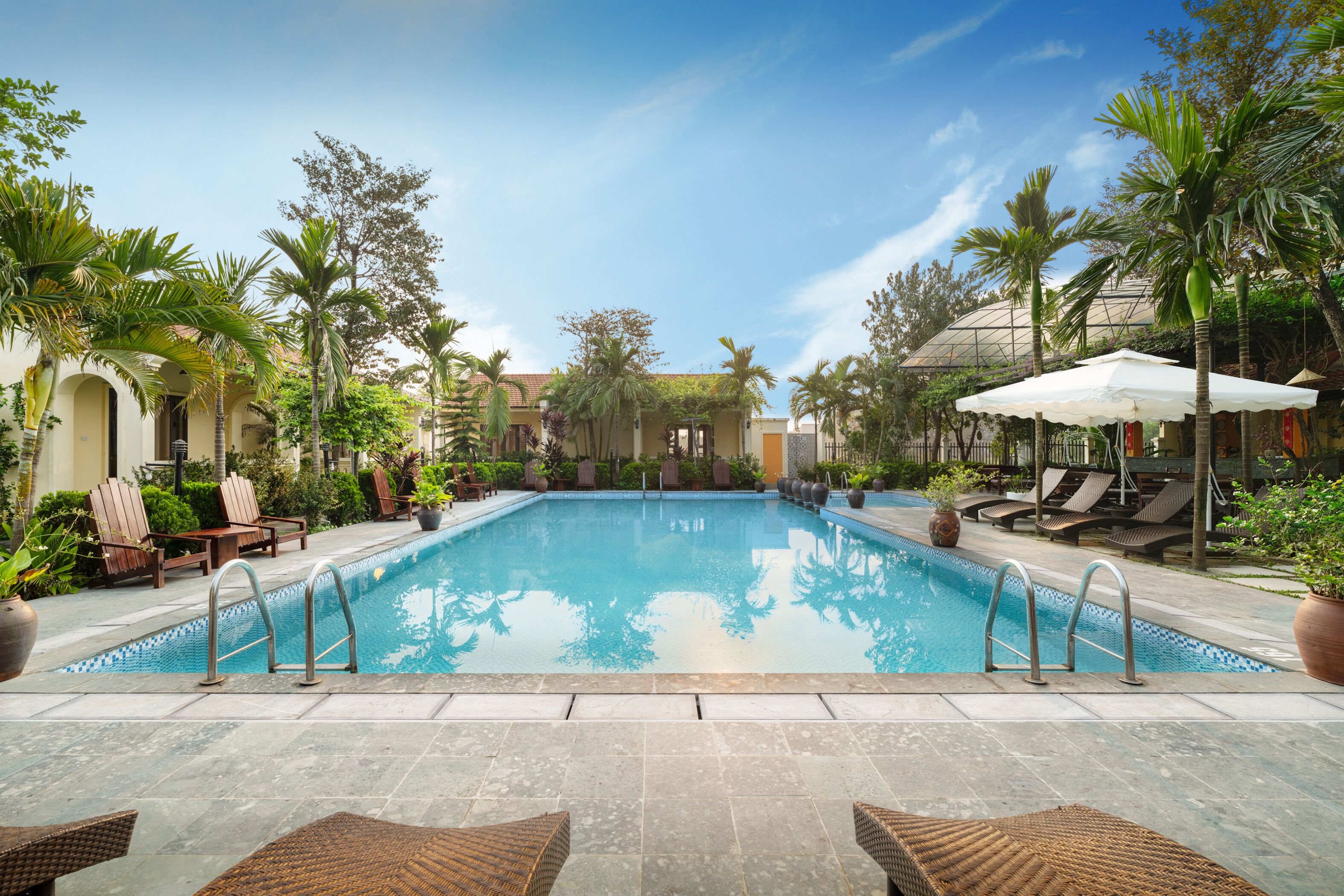 Ein Swimmingpool mit Liegestühlen und Sonnenschirmen, umgeben von Palmen und Kübelpflanzen.