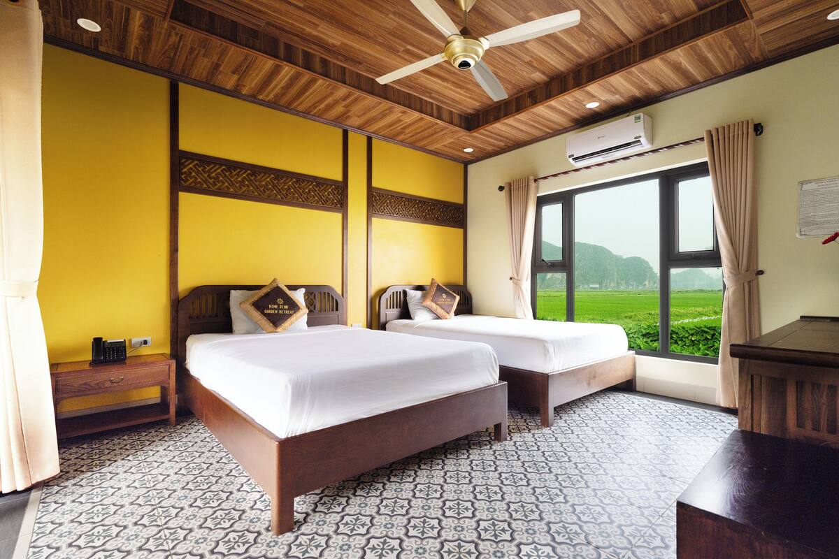 Quarto de hotel com duas camas, ventoinha no teto, ar condicionado e uma janela grande com vista para áreas verdes.