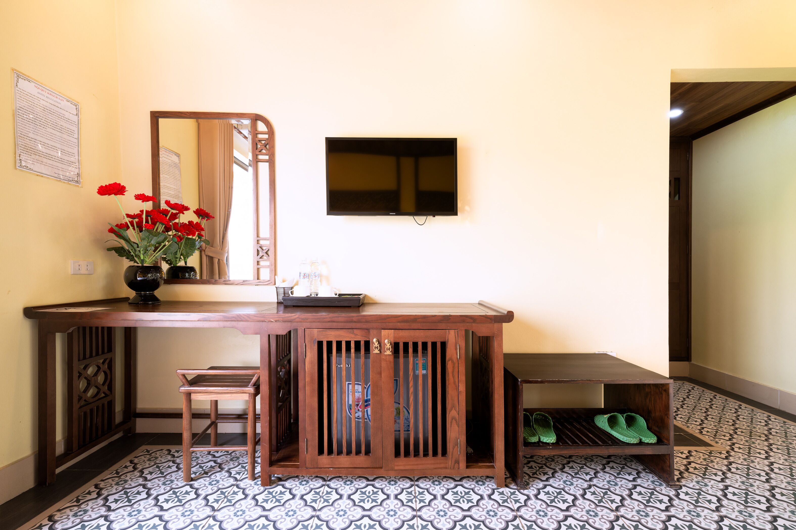 A wooden desk with a mirror, a TV, and a vase of red flowers.
