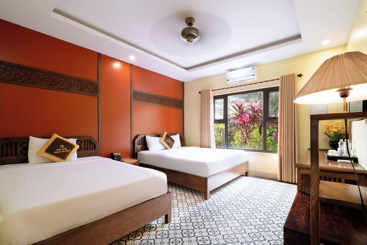 Quarto de hotel com duas camas, uma cabeceira de madeira, piso estampado, janela com cortinas e uma escrivaninha com abajur.
