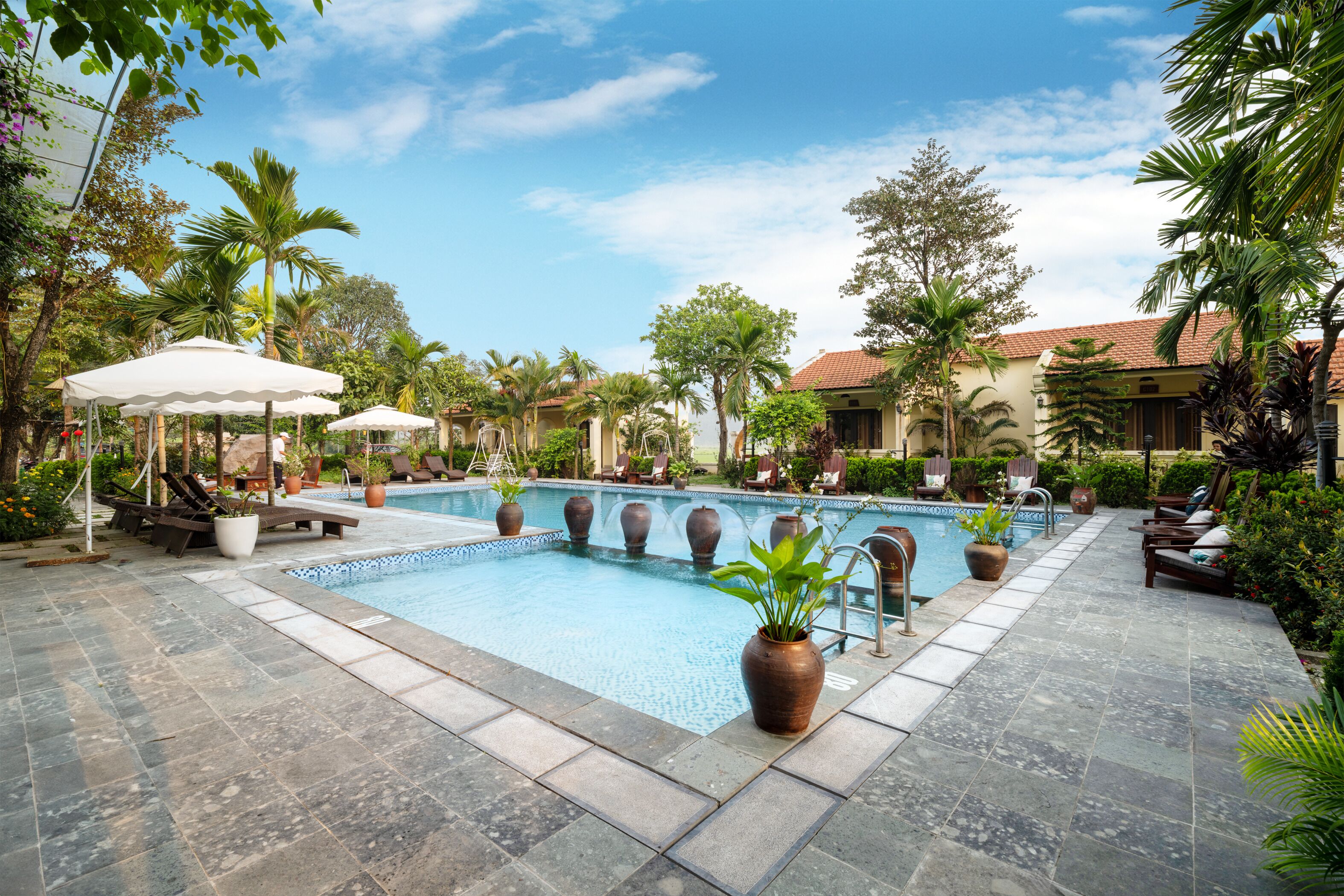 A swimming pool with lounge chairs and umbrellas, surrounded by potted plants and palm trees.