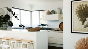 A modern kitchen with white cabinetry, a central island with stools, a large window, and various decorative items. - Pacific Blue by Contemporary Hotels (PALM BEACH)