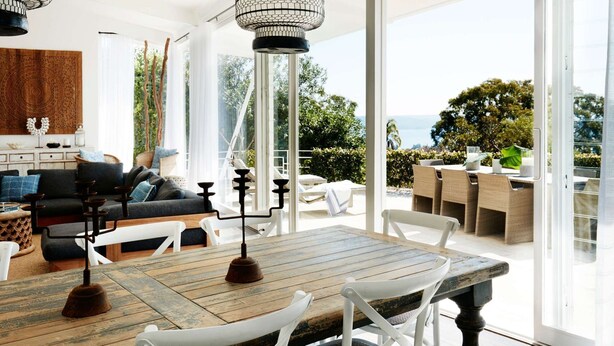 A spacious living area with a large wooden table, white chairs, and a black sofa. There is a view of the outdoors through large windows. - Pacific Blue by Contemporary Hotels (PALM BEACH)