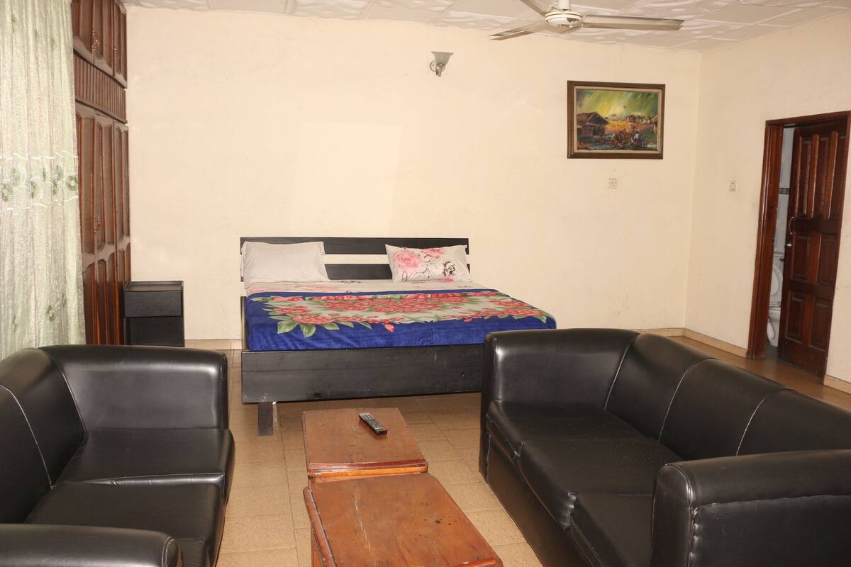 Un dormitorio con una cama, dos sofás de cuero negro, una mesa de centro de madera, un ventilador de techo y un cuadro en la pared.