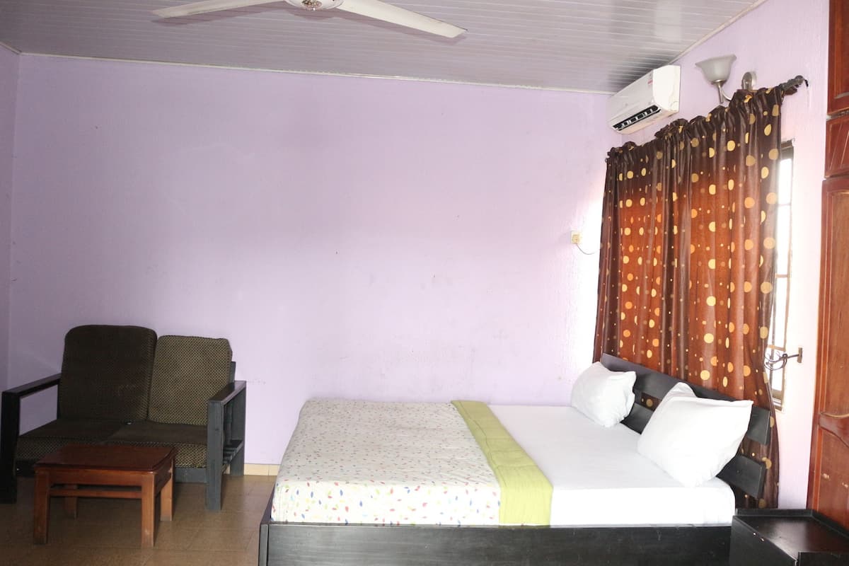 Una habitación con una cama, una silla, una mesita, un ventilador de techo y una ventana con cortinas.