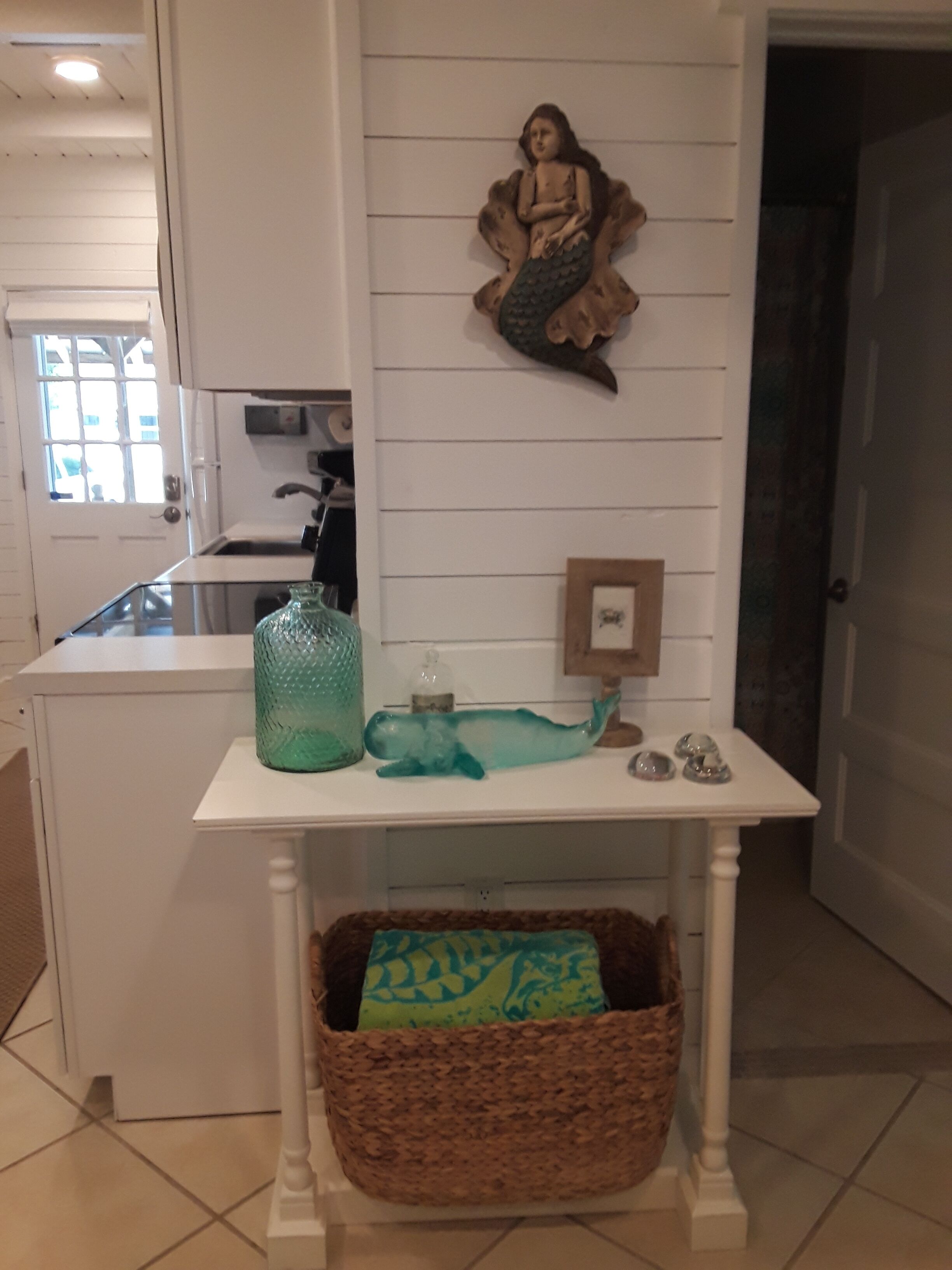Uma cozinha com bancada branca, uma sereia decorativa, um vaso de vidro verde e uma cesta tramada.