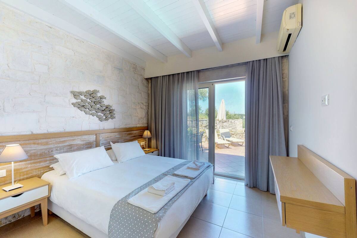 A hotel room with a large bed, a bedside table with a lamp, a wall-mounted air conditioner, and a view of the outdoors through a glass door.