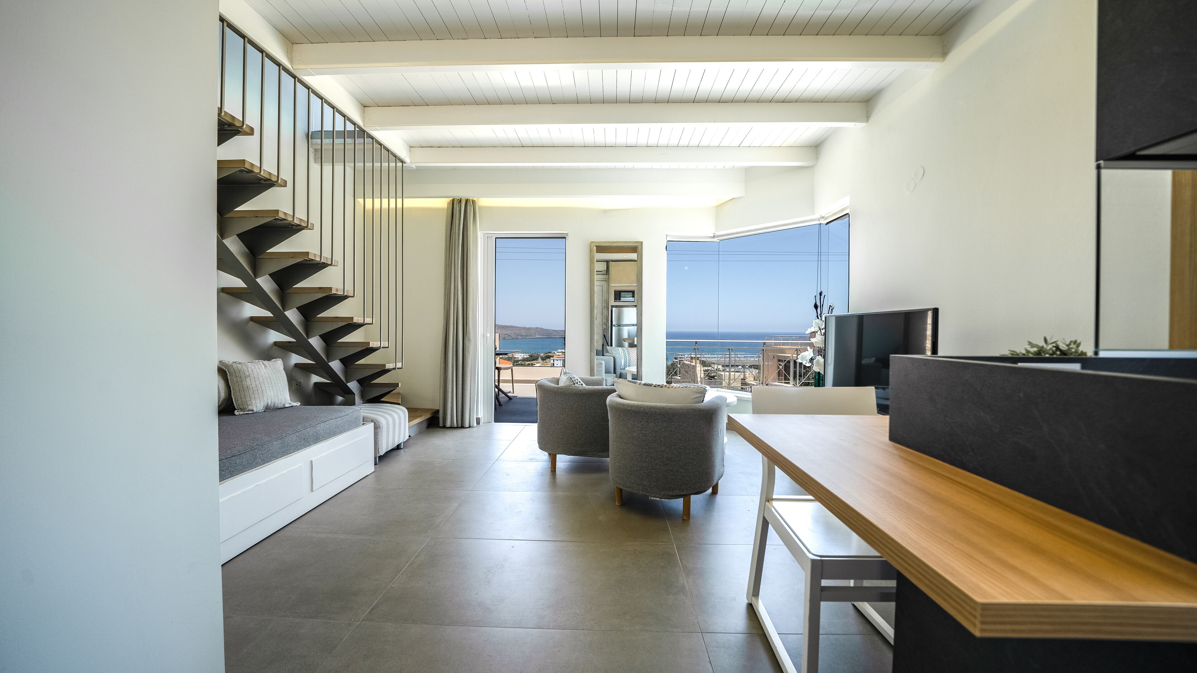 A modern living room with a staircase, a dining area, and a view of the sea.