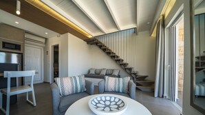 A modern living room with a round white table, a grey sofa with patterned cushions, a wooden chair, and a staircase leading to an upper level. - Lofos Village Chania (Chania)