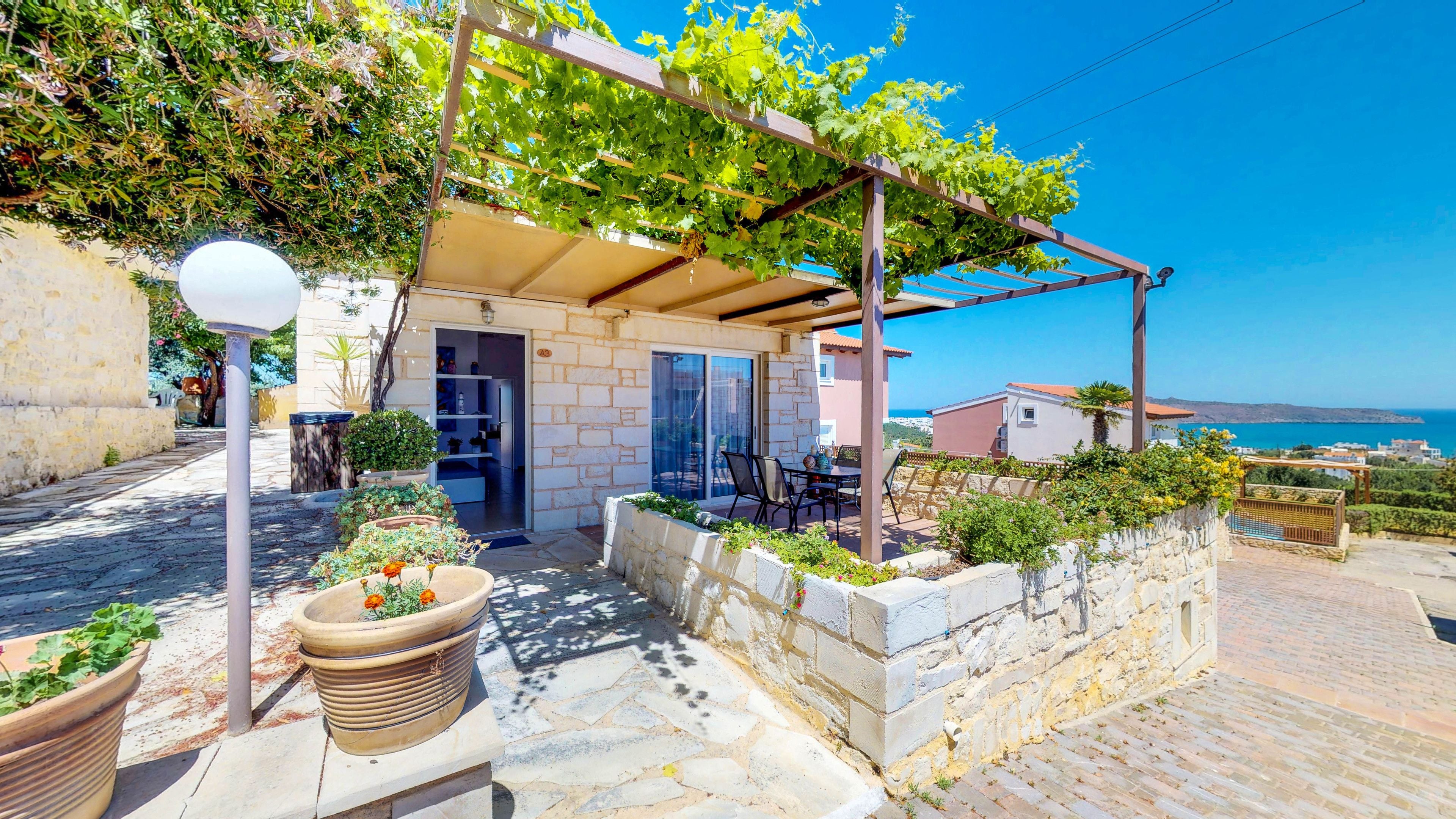 A covered outdoor seating area with a stone wall, potted plants, and a view of the sea.