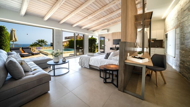 A modern hotel room with a large bed, a sofa, a dining area, and a view of the pool and ocean. - Lofos Village Chania (Chania)