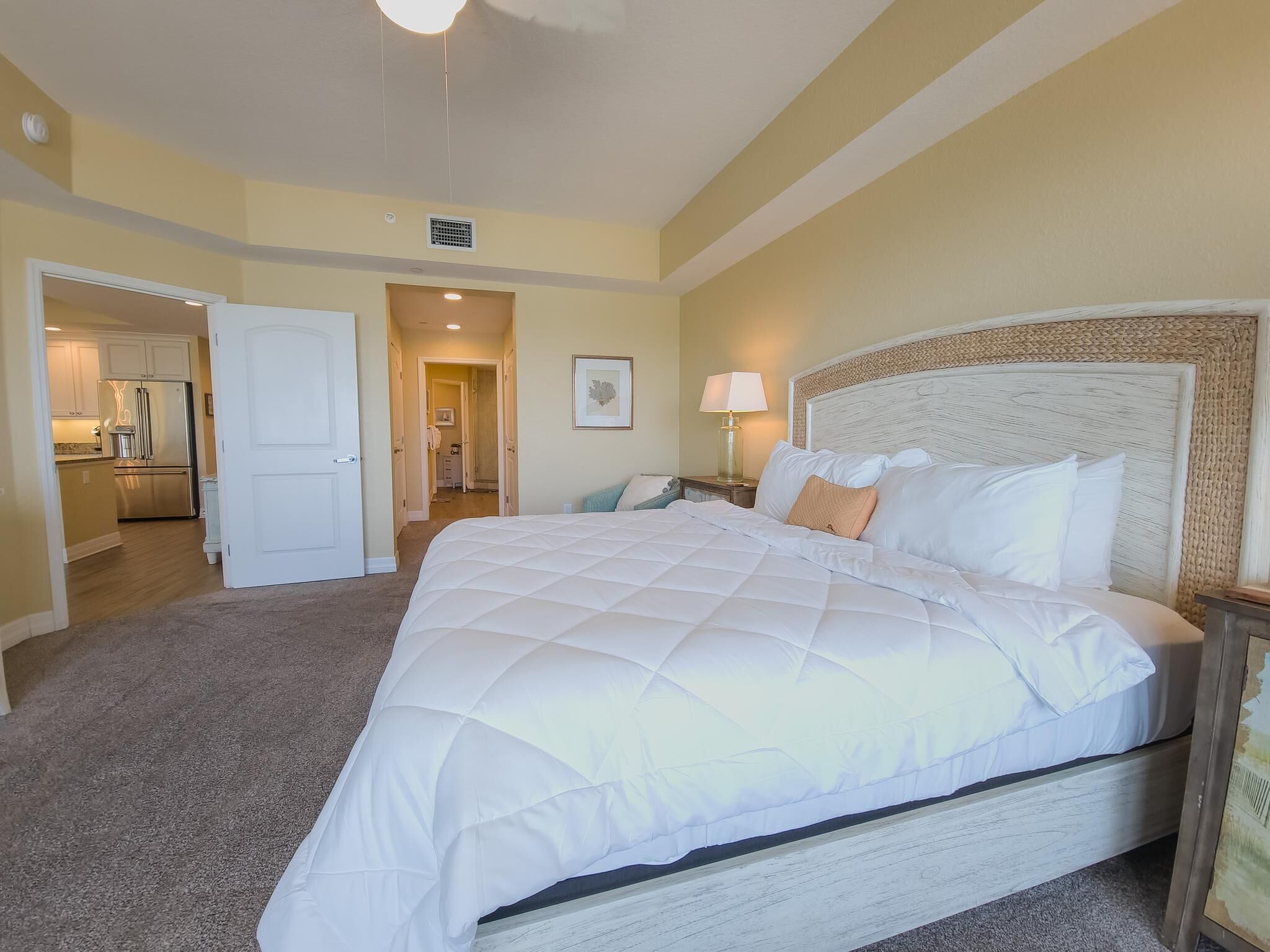 A hotel room with a large bed, a nightstand, a lamp, and a view of the kitchen and bathroom through an open door.