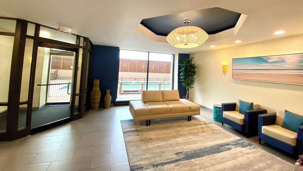 A modern living room with a beige sofa, a blue and beige sectional, a patterned rug, a large window with a view of a balcony, and a ceiling light fixture. - Luxury Oceanfront Condo  (Long Beach)