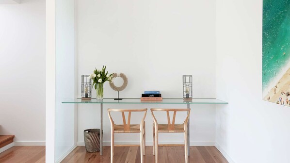 Un bureau à plateau en verre, accompagné de deux chaises en osier, un vase avec des fleurs, un livre intitulé « Regarde ! » et un luminaire fixé au mur.