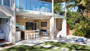 Une maison moderne à deux étages dotée d’un balcon en verre, d’une cuisine avec un îlot central et d’un patio aménagé avec des sièges d’extérieur.