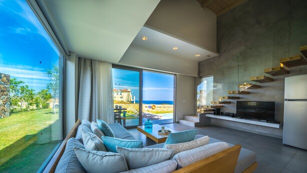 Una sala de estar moderna con un sofá grande, una mesa de centro y un televisor. Se ve el océano y un patio al exterior.