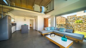 Una cocina moderna con refrigerador empotrado, un comedor con mesa blanca y cristalería azul, y una sala de estar con un amplio sofá modular y una pared de piedra.