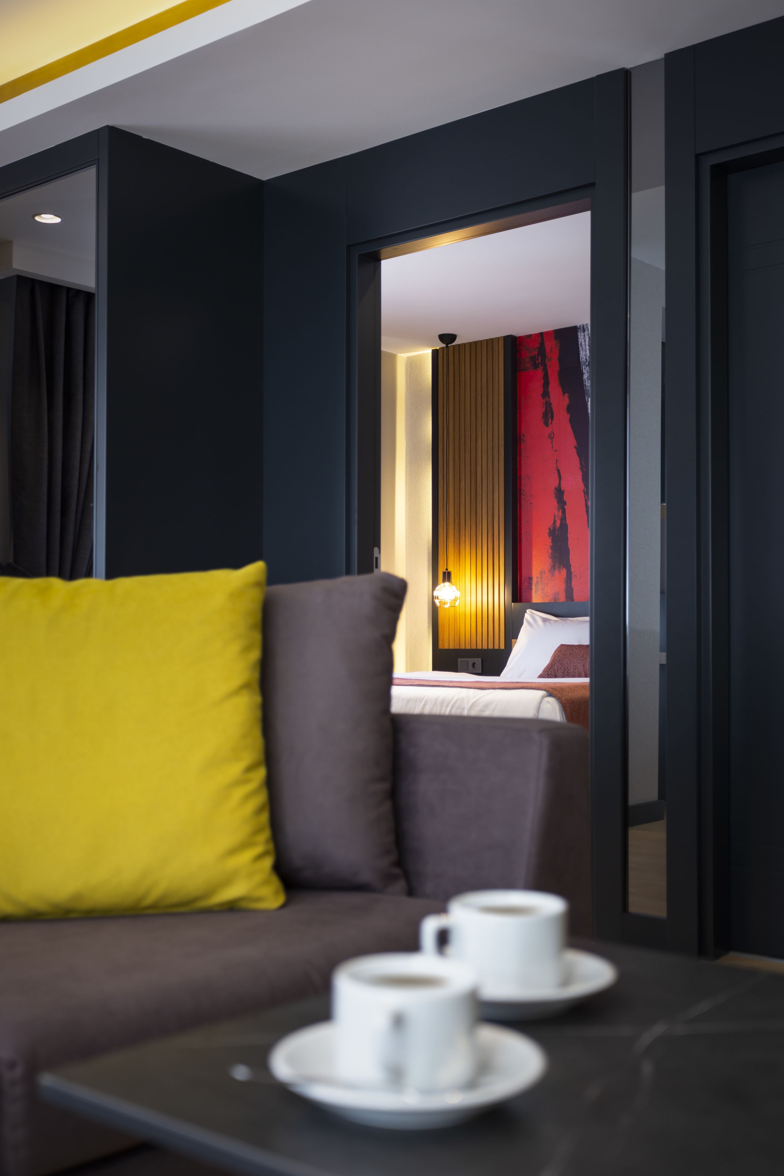 Une chambre d’hôtel moderne avec un canapé de couleur sombre, une table basse sur laquelle reposent deux tasses, et un lit orné d’un tableau abstrait rouge accroché au mur.