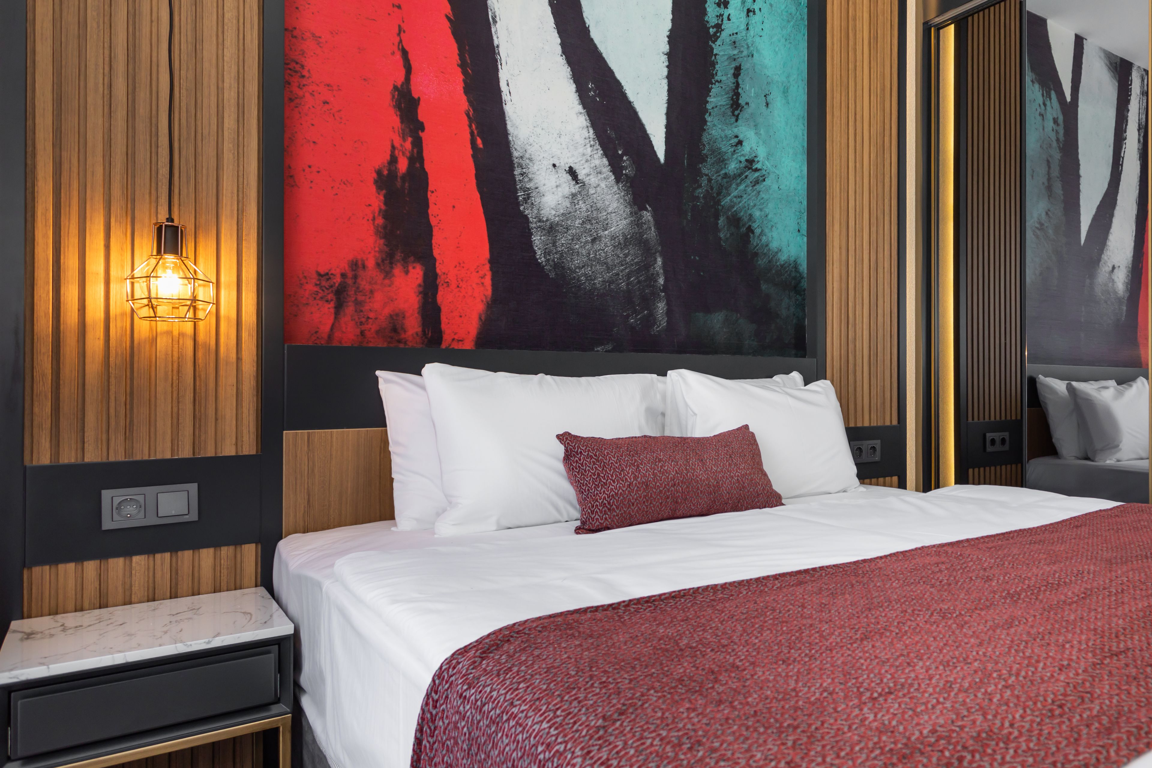 A modern hotel room with a large bed, white bedding, a red throw pillow, a nightstand with a lamp, and a large abstract painting on the wall.