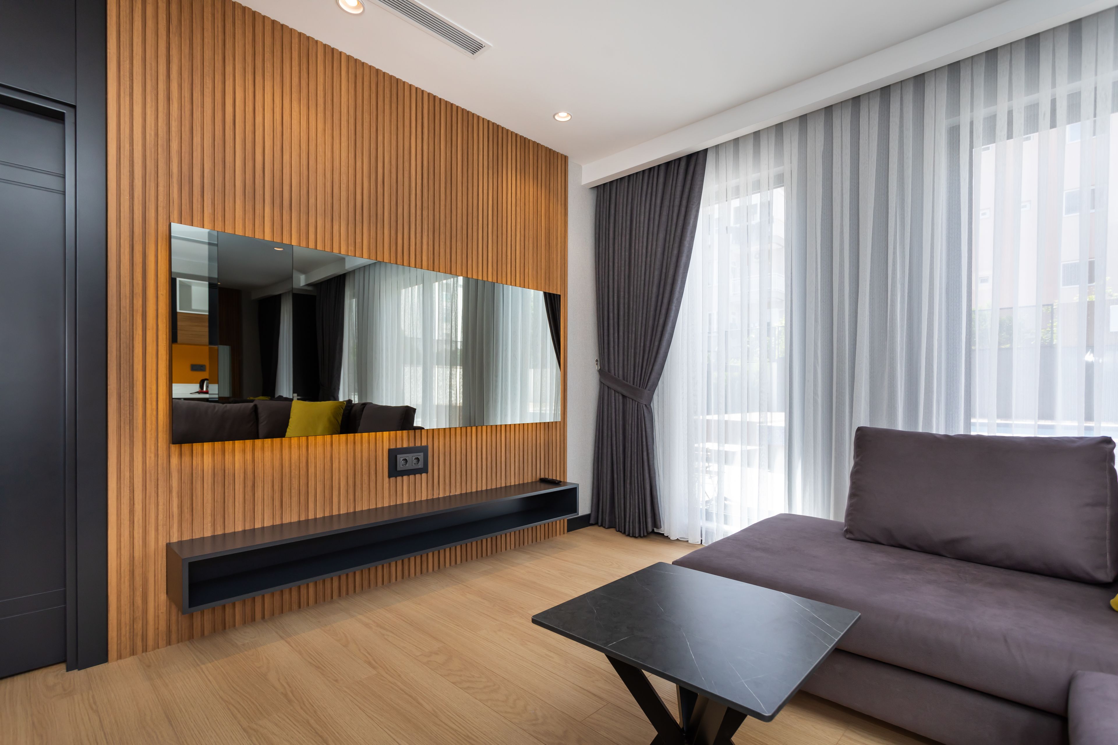 A modern living room with a wooden panel wall, a large mirror, a sofa, a coffee table, and a TV.