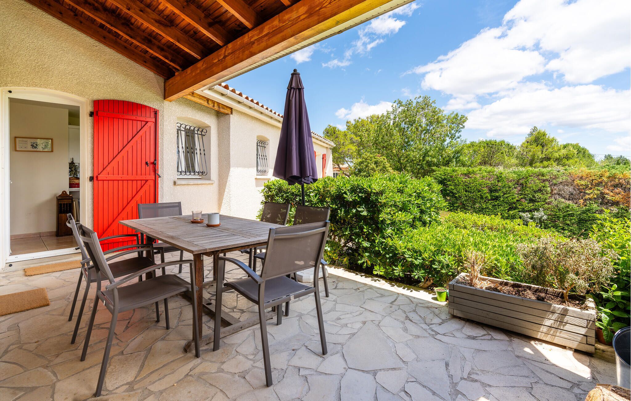 Eine Außenpatio mit Tisch und Stühlen, eine rote Tür und Blick auf einen üppigen Garten.